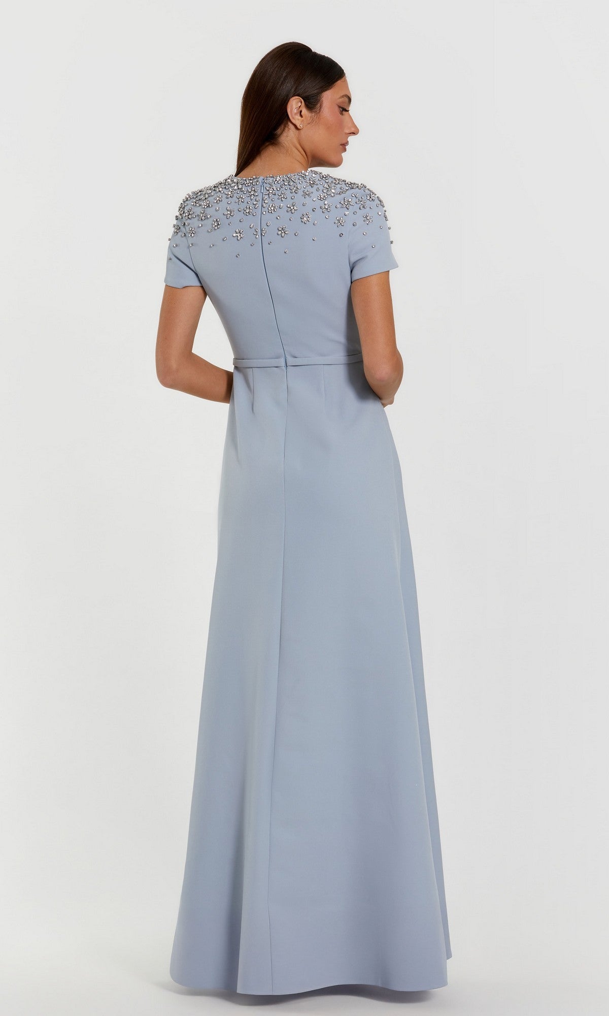 A woman stands with her back turned, wearing the Mac Duggal 50038—a light blue, floor-length crepe trumpet gown featuring short sleeves and embellished detailing on the upper back and shoulders.